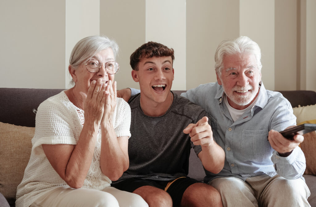 Enthusiastic multigenerational family fans watching Grandma watch a meaningful mother's day gift sitting together on sofa, happy grandparents and young grandson expressing joy