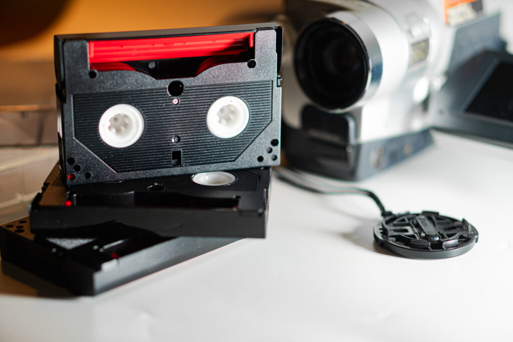 A close-up of VHS tapes stacked with a vintage camcorder in the background. The tapes are black with a red label, showcasing the classic design of video cassettes. The camcorder is silver, indicating an older model, adding to the nostalgic feel of the scene.